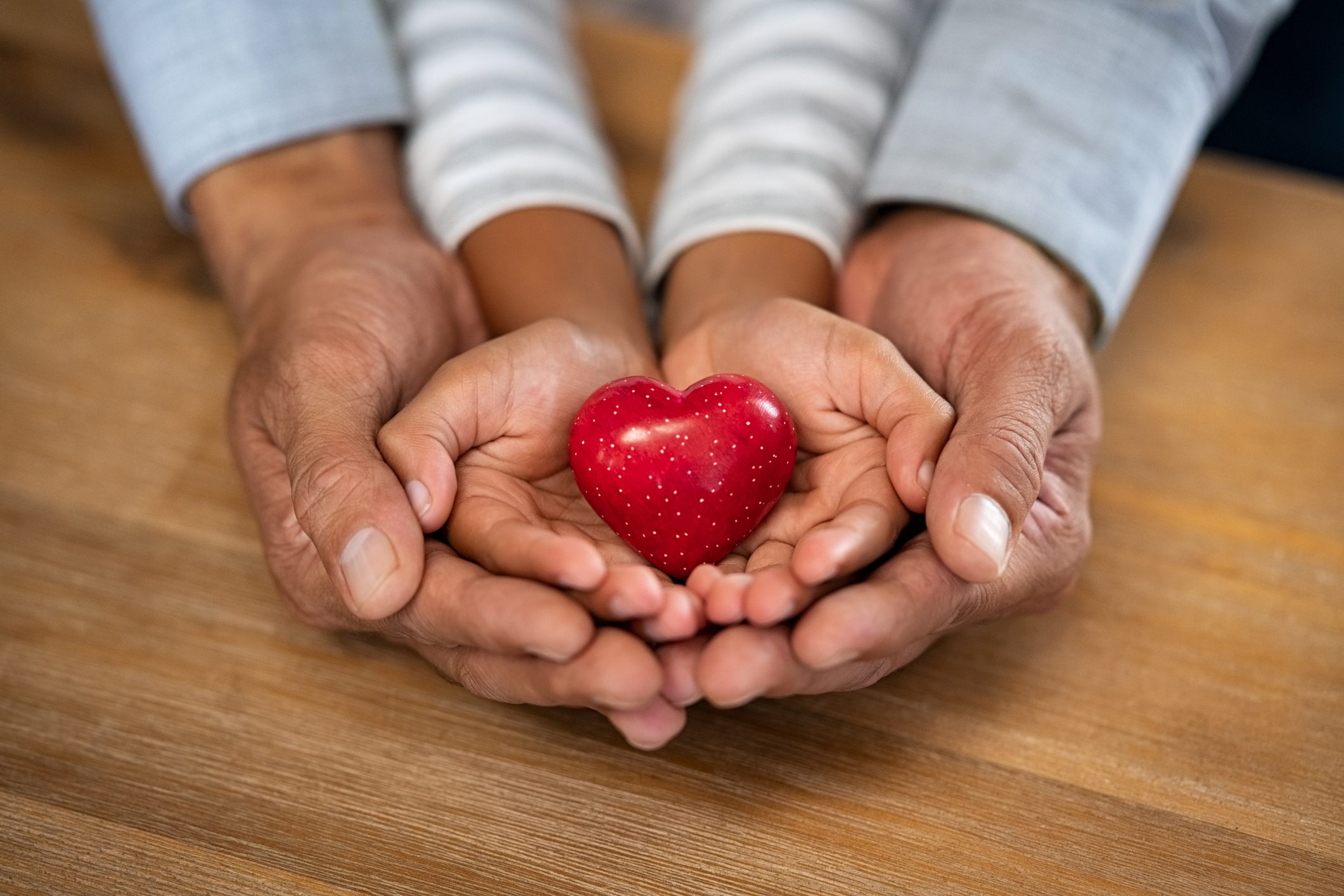 a pair of adult hands and child hands hold a small red heart figurine. 