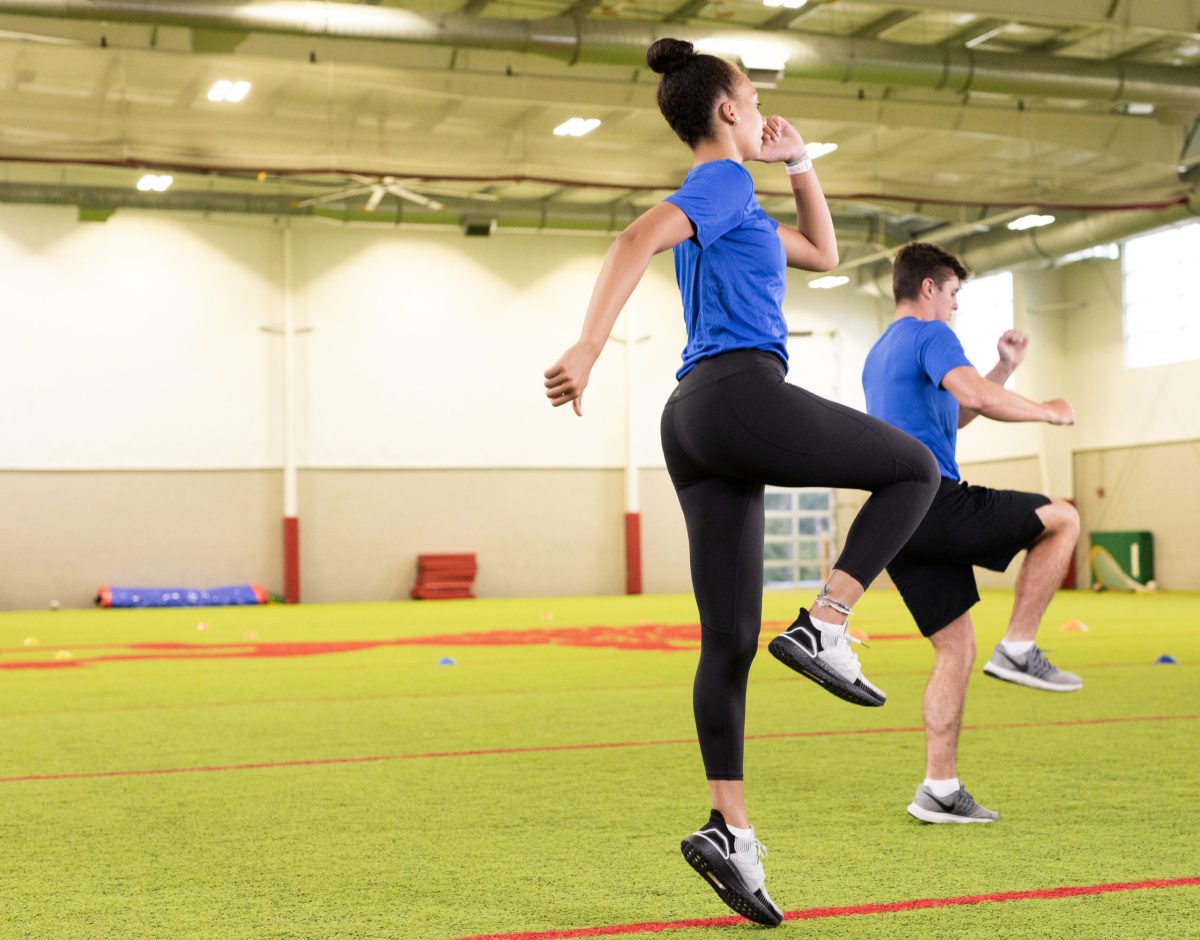 Two student athletes perform a high knee jump exercise in an indoor facility.
