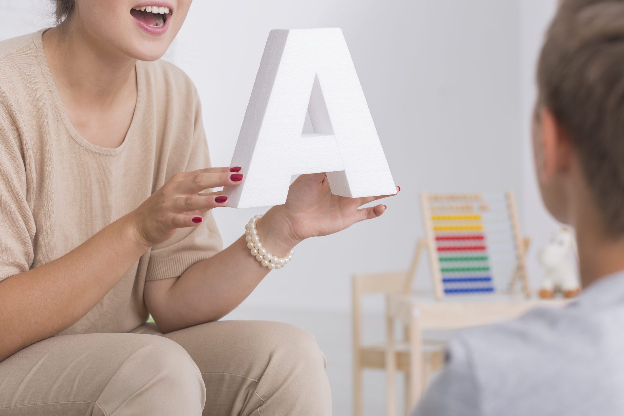 Speech therapist in beige costume holds letter A and speaks to kid during class session.