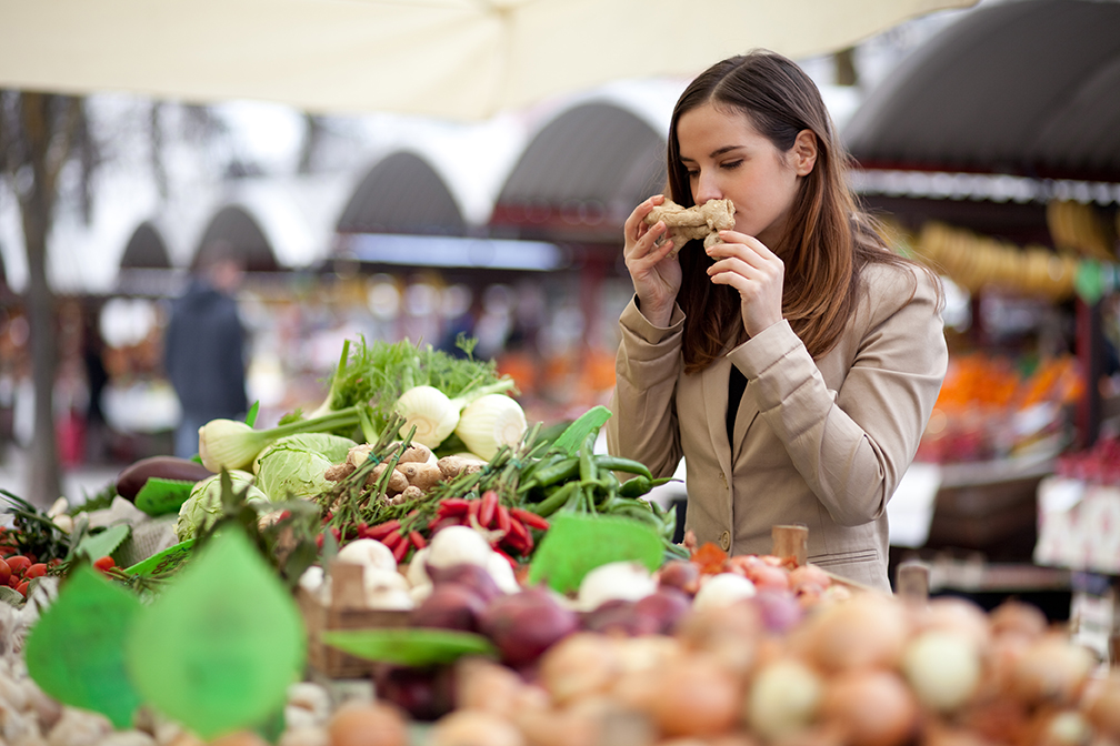 Brown haired female smelling ginger root at farmers market in front of colorful vegetable stand.