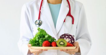 Dietitian in white lab coat with red stethoscope around neck holding plate of fresh fruits and vegetables
