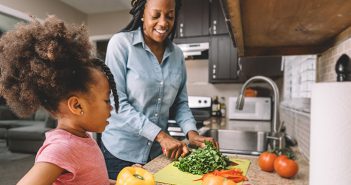 African American mother and daughter cutting vegetables together in kitchen.