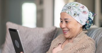Cancer Support Services Remain Available During Pandemic A senior adult woman of Chinese descent has cancer. The happy woman is using a digital tablet to video message friends and family. She is spending time at home. The woman is smiling at the screen. She is wearing a bandana to hide her hair loss from chemotherapy treatment.