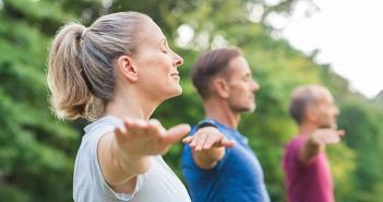 Group of senior people with closed eyes stretching arms at park. Happy mature people doing yoga exercise outdoor on a bright morning. Yoga class with woman and men doing breath exercising with stretched arms.