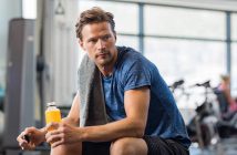 Handsome young man in sportswear holding bottle of fresh orange juice while resting at gym.Thoughtful fit man sitting alone holding a bottle of energy drink. Guy take break after fitness exercise on bench.