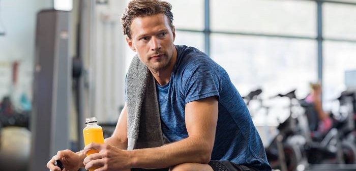 Handsome young man in sportswear holding bottle of fresh orange juice while resting at gym.Thoughtful fit man sitting alone holding a bottle of energy drink. Guy take break after fitness exercise on bench.